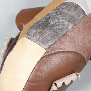 May include: Close-up of a patchwork leather item featuring various shades of brown, tan, and silver. The design incorporates geometric shapes and textures, with visible stitching and a raw edge detail. The item appears to be handcrafted.