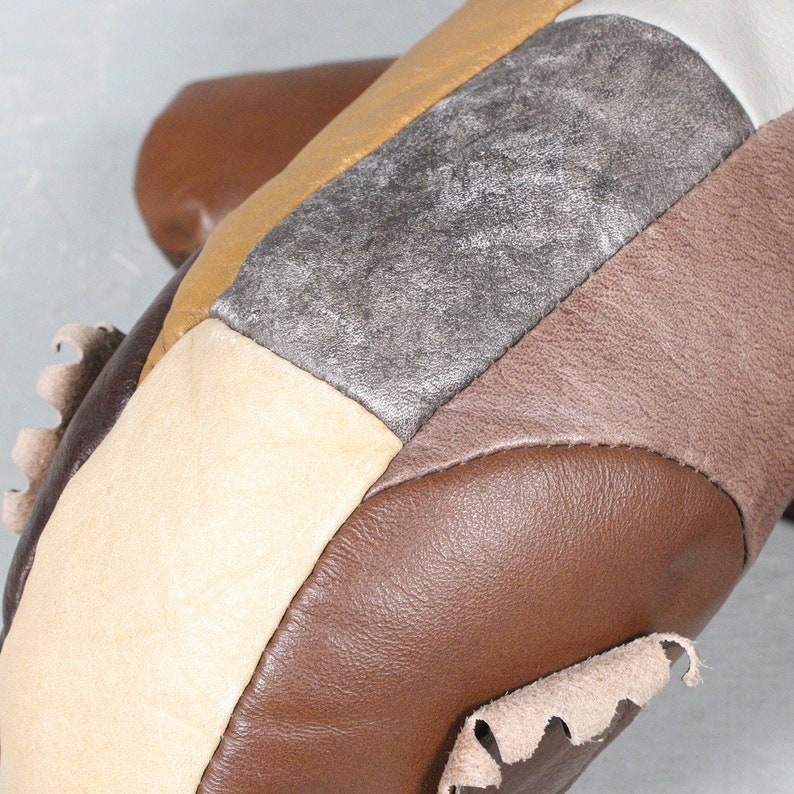 May include: Close-up of a patchwork leather item featuring various shades of brown, tan, and silver. The design incorporates geometric shapes and textures, with visible stitching and a raw edge detail. The item appears to be handcrafted.