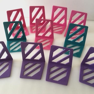 Puede incluir: Colección de sujetalibros geométricos coloridos en tonos rosa, turquesa y morado. Cada sujetalibros presenta una forma cuadrada con cortes diagonales, creando un diseño moderno y elegante. Los sujetalibros están dispuestos sobre una superficie blanca.