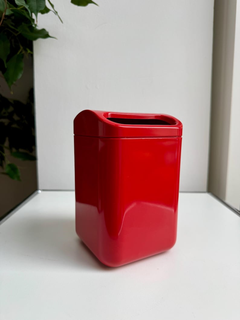 Puede incluir: Un cubo de basura rojo brillante, de forma rectangular con una parte superior curvada y una abertura rectangular. El cubo est&aacute; sobre una superficie blanca, con una pared blanca y una planta verde en el fondo. Dimensiones no visibles.