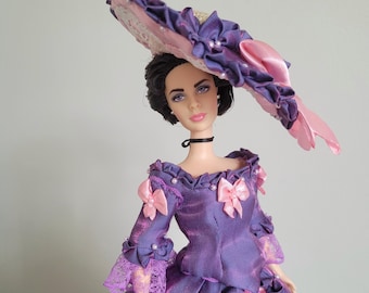 May include: A doll with dark hair wearing a purple and pink gown with ruffles, lace trim, and small white beads. The doll wears a wide-brimmed hat decorated with purple and pink fabric ruffles and a black choker necklace around its neck.