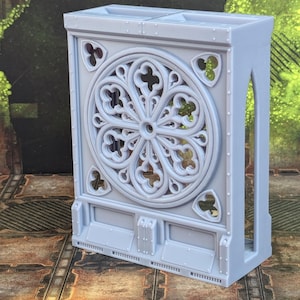 May include: A gray 3D printed miniature wall with a large, detailed, circular window featuring a gothic design. The wall has a flat, rectangular base and a tall, narrow section on the right side.