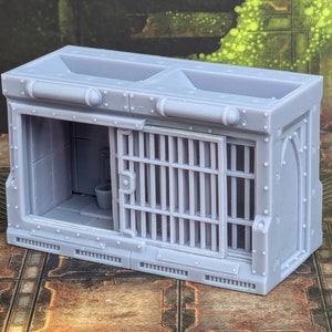 Op de afbeelding: Een grijs, plastic model van een gevangeniscel met een traliedeur en een toilet erin. De cel heeft een klein raam en een metalen dak.