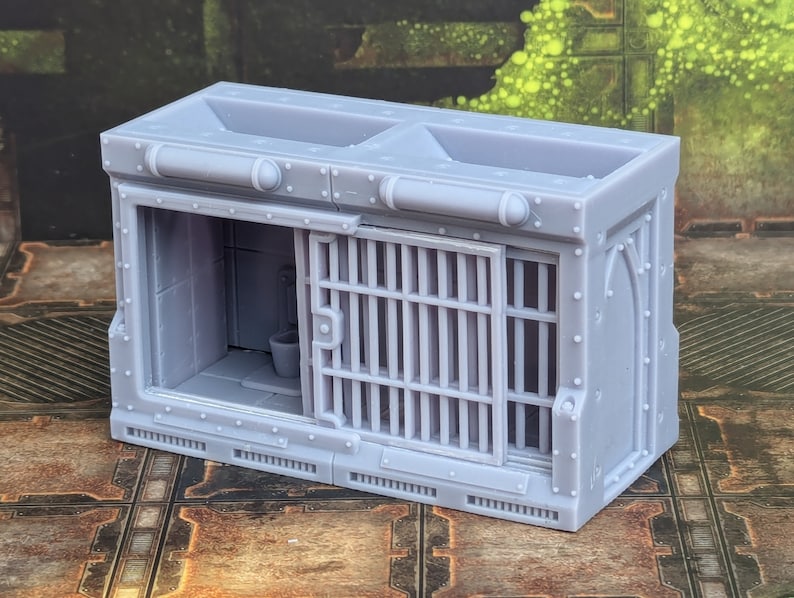 Op de afbeelding: Een grijs, plastic model van een gevangeniscel met een traliedeur en een toilet erin. De cel heeft een klein raam en een metalen dak.