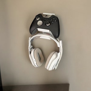 Op de afbeelding: Een witte wandbeugel met een zwarte videogamecontroller en een witte koptelefoon die eraan hangen.