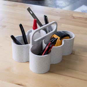 Op de afbeelding: Een witte, multi-compartimenten organizer met een handvat, ontworpen om verschillende gereedschappen en benodigdheden te bewaren. Hij heeft een rode fles lijm, zwarte pennen, een gele rolmaat en een zilveren stanleymes.