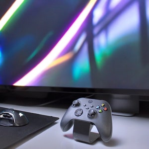 May include: A black Xbox controller with a stand on a white surface. The controller has a black and green colour scheme with a black and white mouse mat and a black mouse in the background.