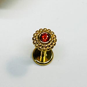 Puede incluir: Un pendiente labial dorado con un centro de piedra roja. El pendiente tiene un diseño floral alrededor de la gema. La base del pendiente es redonda y plana. El fondo es blanco.