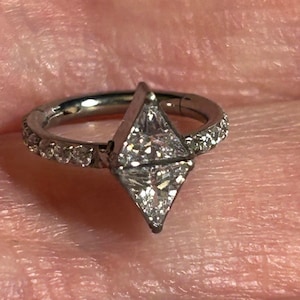 May include: A silver hoop earring with a clear, faceted, triangular gemstone set in the center. The hoop is adorned with a row of small, clear gemstones.