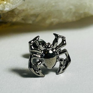Puede incluir: Un pendiente de botón plateado con forma de araña. El pendiente presenta un diseño detallado con ocho patas y un cuerpo redondeado. La araña está centrada en una superficie blanca, con un fondo borroso.