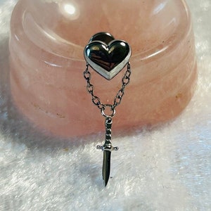May include: A silver heart-shaped earring with a dangling silver dagger charm attached to a silver chain.
