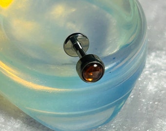 May include: A small, silver-colored stud earring with a round, amber-colored gemstone. The earring is set against a blurred, opalescent background with shades of blue, white, and yellow.