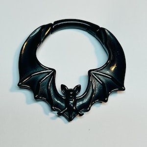 May include: A black, circular ear gauge with a bat design. The gauge features a detailed bat silhouette with outstretched wings and a small bat head in the center. The gauge is made of a shiny, dark material.