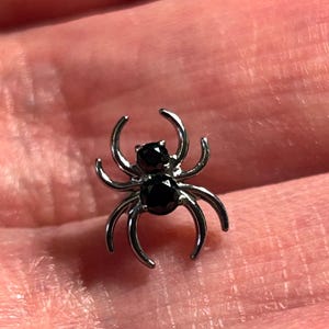 May include: A small, silver spider charm with eight curved legs and two black gemstone eyes. This decorative spider charm is a jewellery accessory.