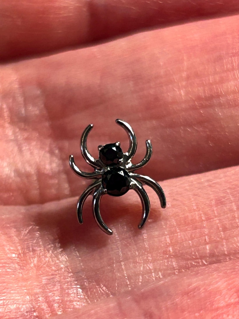 May include: A small, silver spider charm with eight curved legs and two black gemstone eyes. This decorative spider charm is a jewellery accessory.