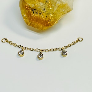 May include: A gold-toned chain connector featuring three small, round, clear bezel-set stones hanging from the links. The chain has a small circular jump ring at each end, and a large, translucent, faceted yellow crystal sits above the chain.