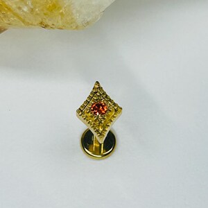 May include: A gold-colored, diamond-shaped earring with a small, round, orange gemstone in the center. The earring has a textured surface and a flat, circular base. The design is intricate and detailed, showcasing a delicate craftsmanship.