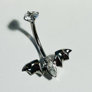 Puede incluir: Un piercing para el ombligo plateado con un diseño de murciélago. La barra curva tiene una gema grande y transparente de talla redonda en la parte superior y una gema en forma de pera en el centro del cuerpo del murciélago. Las alas del murciélago están detalladas.