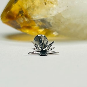 Op de afbeelding: Een zilverkleurige oorbel met een sunburst-ontwerp en een kleine, zwarte edelsteen in het midden. De oorbel heeft een pin-achterkant en is geplaatst tegen een witte achtergrond met een vage, geel-oranje kristal op de achtergrond.