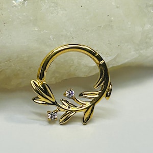 May include: Gold-colored septum ring featuring a leaf design with two small, square-cut, clear gemstones. The ring is circular with a hinged closure. The leaf design is on the lower half of the ring.