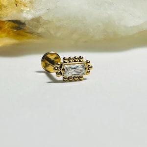 May include: A gold-colored stud earring with a rectangular, clear gemstone at the center. The gemstone is surrounded by small, round gold beads. The earring is set against a white background.
