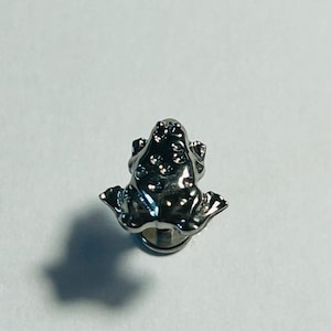 Può includere: Una spilla da bavero a forma di rana color argento. La spilla presenta un design dettagliato con piccoli abbellimenti rotondi sulla schiena della rana. La rana è raffigurata in posizione seduta con le zampe divaricate. La spilla ha un perno sul retro per il fissaggio.