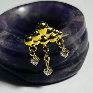 May include: A gold-colored cloud-shaped charm with three dangling cubic zirconia stones. The charm is set against a dark purple, textured background. The cloud has a textured surface and is made of a shiny, reflective material.