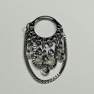 May include: A silver-toned septum ring with a dangling chain and crystal accents. The ring features a circular top with an oval-shaped chain design and multiple dangling crystals. The jewelry is designed for nose piercings.