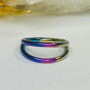 Puede incluir: Un anillo de doble aro con un acabado iridiscente vibrante. El anillo presenta un espectro de colores, incluyendo azules, púrpuras y dorados. La joya se encuentra sobre un fondo neutro, resaltando su diseño colorido.