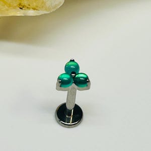 Pode incluir: Uma joia corporal tipo labret em tom prateado com um grupo de três pequenas contas redondas, lisas e de um verde-azulado vibrante, dispostas em forma triangular no topo de uma haste reta com uma base circular plana.