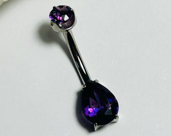 Peut inclure: Un bijou de corps en forme de barre courbe argentée, orné d'une petite pierre précieuse ronde violette sur le dessus et d'une plus grande pierre violette en forme de poire en bas, fixées par des griffes sur une tige métallique polie.