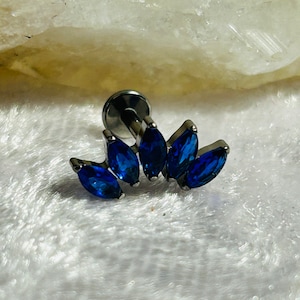 Könnte beinhalten: Ein silberner Körperschmuck mit einer Reihe von fünf blauen, marquiseförmigen Steinen. Die Steine sind in einer gebogenen Form gesetzt, wodurch ein Halbmond-Design entsteht.