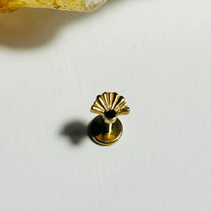 Könnte beinhalten: Ein kleiner, goldfarbener Ohrstecker mit einem fächerförmigen Design. Der Ohrring hat eine flache, kreisförmige Basis und eine dekorative Oberseite mit mehreren gebogenen Segmenten. Der Ohrring ist vor einem weißen Hintergrund.