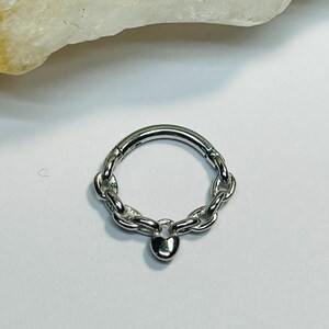 May include: A silver nose ring featuring a chain design and a small heart pendant. The ring is circular with a hinged closure. The heart pendant is suspended from the chain's centre. The background is white.