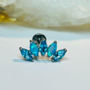 Puede incluir: Un pendiente de plata con cinco piedras de color azul claro en forma de marquesa dispuestas en forma de abanico. Las piedras tienen un ligero efecto iridiscente. La joya está diseñada para piercing.