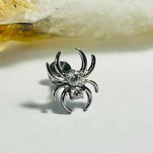 Puede incluir: Un pendiente de botón plateado con forma de araña con dos gemas claras y brillantes. La araña tiene patas curvas y está sobre un fondo blanco. La joya está diseñada para ser usada como piercing en la oreja.