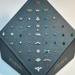Può includere: Un espositore nero testurizzato con il logo ZURA, che mostra una collezione di vari gioielli da corpo in metallo con pietre preziose di diversi colori, forme e design a catenella pendente disposti in una formazione a piramide.
