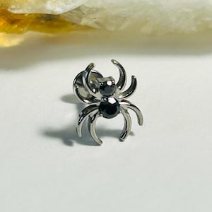 May include: A silver spider stud earring with two black gemstones. The spider design features eight curved legs and a small post. The earring is set against a white background.