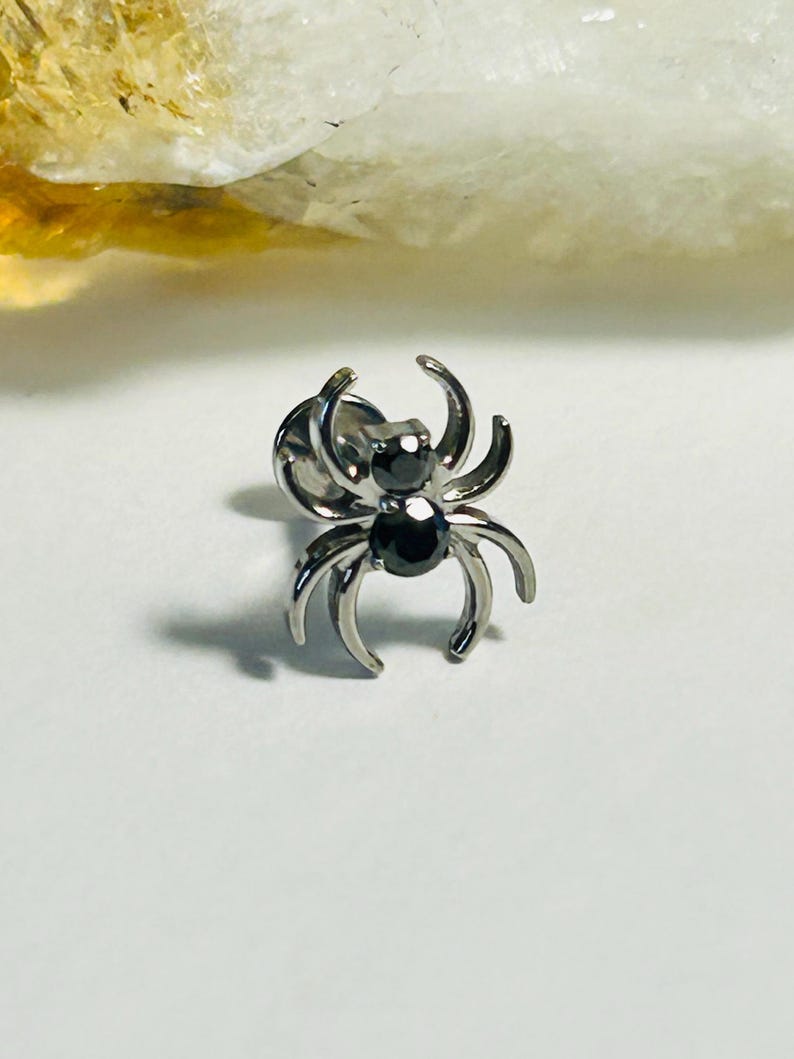 May include: A silver spider stud earring with two black gemstones. The spider design features eight curved legs and a small post. The earring is set against a white background.