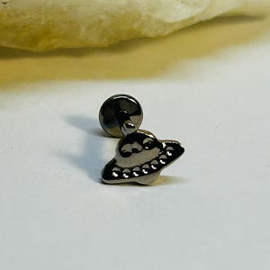 Puede incluir: Un pendiente de plata con un pequeño y detallado diseño de OVNI. El pendiente tiene un poste y una parte trasera plana para un uso seguro. El OVNI tiene una forma de platillo clásico con pequeñas luces espaciadas uniformemente.