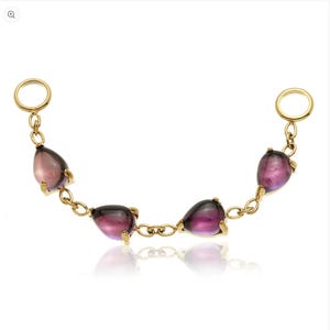 May include: A gold chain featuring five teardrop-shaped, purple and clear gemstones. The gemstones are set in gold prongs and linked by a delicate chain. Two gold rings are at each end of the chain.