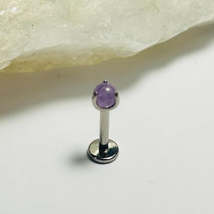 Può includere: Un piercing per labbra color argento con una piccola gemma rotonda viola chiaro. Il piercing ha una base piatta e circolare e un perno dritto che conduce alla gemma. La gemma è incastonata in una lunetta d'argento.
