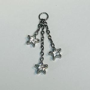 Puede incluir: Charm de tono plateado con tres colgantes de estrella. Cada estrella tiene un centro transparente y facetado. El charm está conectado a un pequeño anillo en la parte superior mediante delicadas cadenas. Las estrellas miden aproximadamente 1,3 cm.