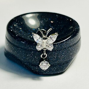 May include: A small, dark blue, speckled jewelry box with a silver butterfly-shaped embellishment. The butterfly has clear, sparkling accents and a dangling, round gem. The box is smooth and rounded.