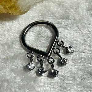 May include: A silver dangle nose ring with a teardrop-shaped base and five dangling clear crystal stones.