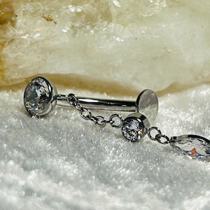 Puede incluir: Un piercing de cuerpo de plata con una cadena y tres piedras de cristal transparentes. El piercing está diseñado para el ombligo u otra parte del cuerpo.