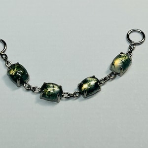 Könnte beinhalten: Ein silberfarbenes Kettenarmband mit fünf grünen und weißen Moosachatsteinen. Die Steine sind rechteckig und durch kleine Kettenglieder verbunden. Das Armband hat an jedem Ende einen Ring zum Befestigen.