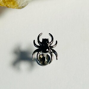May include: A silver spider stud earring with a black body. The spider has eight legs and is set against a white background. The earring is a small, detailed piece of jewellery.