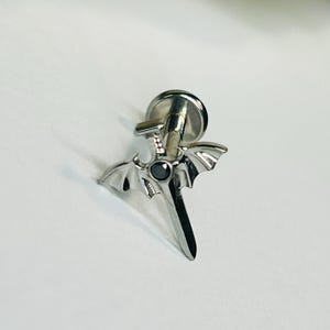 Può includere: Orecchino argentato con un design a spada e una gemma nera. L'orecchino ha un retro piatto e dettagli a forma di ali. Il design è una piccola rappresentazione dettagliata di una spada.