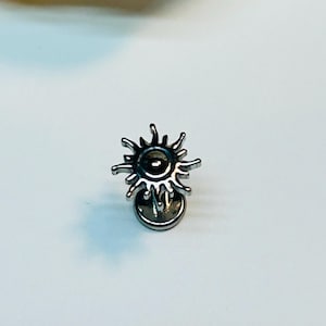 May include: A silver-coloured sun-shaped earring stud. The earring features a detailed sun design with pointed rays extending from a central, dark-coloured sphere. The earring is designed to be worn on the ear.